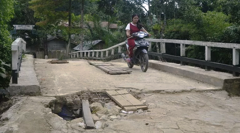 Warga berkendara melalui jembatan sungai Boli yang kondisi lantainya sudah berlubang